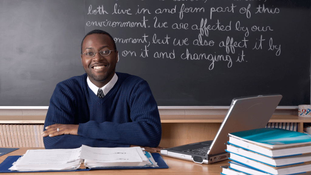 previdência professores - homem negro lecionando, sentado na mesa, com arquivos, livro, computador